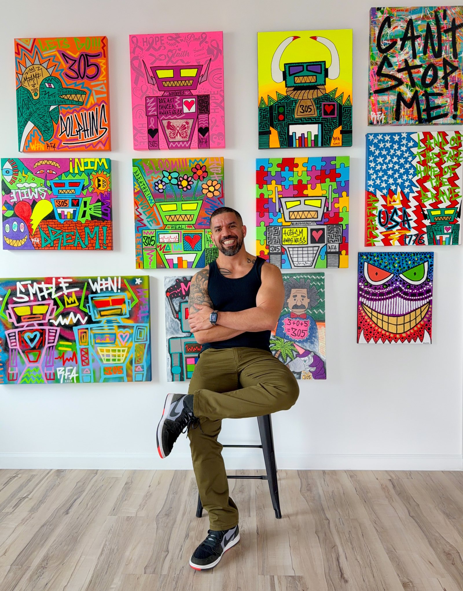 Rick F. Alvarez on a stool in front of a wall hung with twelve of his brightly colored robot paintings.